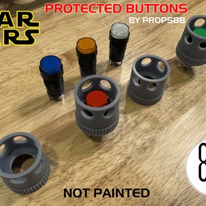 May include: Six gray plastic button covers with different colored buttons inside. The buttons are red, orange, blue, green, white, and clear. The text "PROTECTED BUTTONS BY PROPSS88" is at the top of the image. The text "NOT PAINTED" is at the bottom of the image. A white circle with the text "8P8 PROPS" is in the bottom right corner.