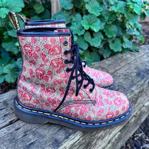 May include: Pair of floral print boots with red roses and green leaves on a tan background. The boots have black laces, black trim, and a black sole with yellow stitching. The boots are made of leather.