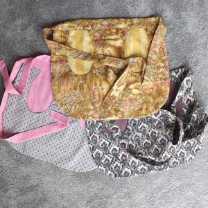Könnte beinhalten: Drei Stoffschürzen in verschiedenen Mustern. Eine ist rosa mit weißen Punkten, eine andere ist braun mit Blumen- und geometrischen Mustern und die dritte ist dunkelbraun mit einem Paisley-Muster.