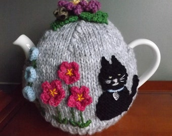 Puede incluir: Una funda de tetera de punto gris con un gato negro, flores rosas y amarillas y una flor morada en la parte superior.