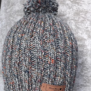 Könnte beinhalten: Graue Strickmütze mit großem Bommel oben. Die Mütze hat eine gerippte Struktur und ist mit orangefarbenen, roten und blauen Flecken gesprenkelt. Ein braunes Lederetikett mit der Aufschrift "Hand Made" ist am Rand angenäht.