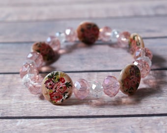 Pode incluir: Uma pulseira de contas facetadas rosa e transparentes, com contas redondas e planas com um desenho floral. O desenho floral apresenta tons de rosa e vermelho. A pulseira é exibida sobre uma superfície de madeira.