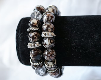 May include: Two beaded bracelets with large, round beads in shades of brown and white. The bracelets feature silver-toned spacers with clear and silver accents. The bracelets are displayed on a black velvet surface.