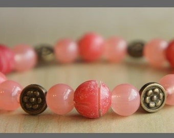 Pode incluir: Uma pulseira de contas rosa com uma mistura de contas redondas e texturizadas. A pulseira apresenta uma variedade de tons de rosa, do claro ao coral profundo, e é acentuada com espaçadores de cor bronze antigo.