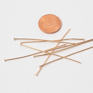 May include: A group of gold head pins scattered on a white surface. A penny is included for scale.