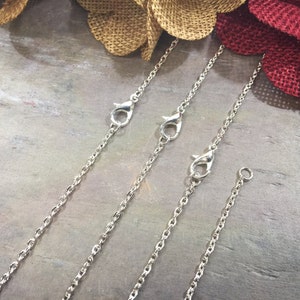 May include: Three silver chains with lobster claw clasps. The chains are on a gray textured surface.
