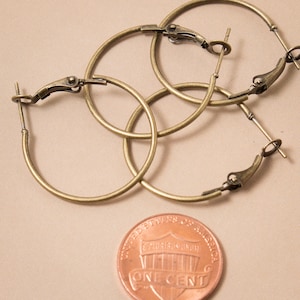 May include: Three pairs of gold-colored hoop earrings with hinged closures. The earrings are circular and have a simple, classic design. A copper penny is next to the earrings for size comparison.