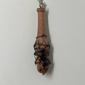 May include: A wooden baseball bat wrapped in barbed wire with a red stain on the wood. The bat has a silver keychain loop attached to the top.