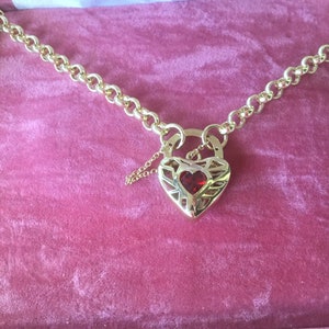 May include: A gold chain necklace with a heart-shaped padlock pendant. The padlock has a red gemstone in the center and intricate detailing.