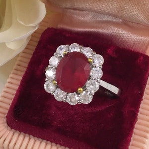 May include: A red oval gemstone ring with a halo of clear round stones. The ring is set in silver and is displayed in a red velvet ring box.