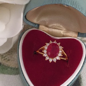 Puede incluir: Un anillo de oro con un gran rubí ovalado rodeado de piedras más pequeñas y transparentes. El anillo está en una caja de anillo de terciopelo rojo.