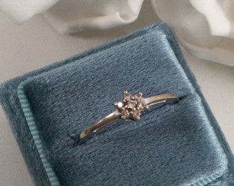 Puede incluir: Un anillo de plata con un grupo de pequeños diamantes engastados en el centro. El anillo se muestra en una caja de anillo de terciopelo azul.