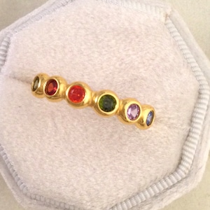May include: A gold band ring with a row of colourful gemstones. The stones are set in a bezel setting and are arranged in a rainbow pattern. The ring is a size 7.