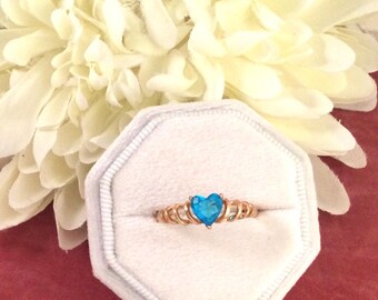 May include: A gold-colored ring with a heart-shaped, bright blue gemstone. The ring is set in a decorative band and is displayed in a white, octagonal ring box. A large, white flower is in the background.