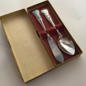 Pode incluir: Um conjunto de talheres vintage em tons de prata, incluindo uma colher, uma faca e uma colher, apresentado numa caixa de cartão com interior vermelho. As pegas dos utensílios apresentam um design detalhado com um retrato e o ano de 1938.