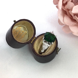 Puede incluir: Un anillo de plata con una gran piedra verde talla esmeralda y piedras blancas más pequeñas, exhibido en un joyero de cuero marrón antiguo abierto con interior dorado. La caja tiene texto en el interior. Una flor rosa está en el fondo.