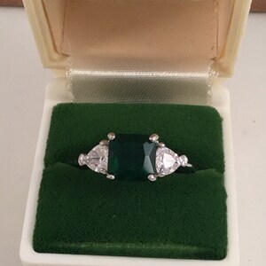 Puede incluir: Un anillo de plata con una gran gema verde de talla esmeralda flanqueada por dos gemas blancas triangulares más pequeñas. El anillo se muestra en un joyero vintage de color crema con interior de terciopelo verde.