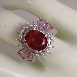 May include: A silver ring featuring a large red oval gemstone surrounded by smaller clear stones. The ring has a halo of pink stones around the centre stone.