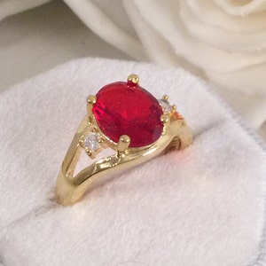 以下が含まれることがあります： 大きな楕円形の赤い宝石と、バンドに2つの小さな透明な宝石が付いた金の指輪です。