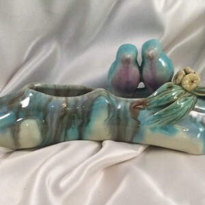 Può includere: Un vaso da fiori in ceramica a forma di ramo d'albero con due uccelli blu appollaiati in cima. Il vaso è smaltato in tonalità di blu, verde e bianco.