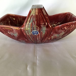 May include: A red and green glazed ceramic planter with a handle. The planter has a geometric design and a blue label that reads "Art Midway Ware".