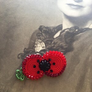 May include: A handmade red poppy brooch with black accents and a green leaf detail. The brooch features two circular red flower heads with a spiral design, embellished with red beads and black dots.