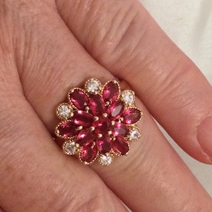 May include: A gold ring with a large red ruby flower design surrounded by smaller white diamonds.