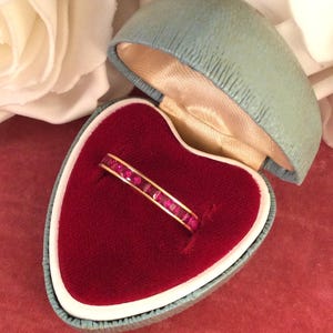 Puede incluir: Un anillo de oro con una banda de piedras preciosas rojas vibrantes, talladas en forma cuadrada. El anillo se muestra en un joyero vintage en forma de corazón con interior de terciopelo rojo y exterior azul claro.