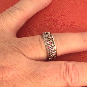 May include: Silver-toned ring with an openwork design featuring small, vibrant pink gemstones. The ring is wide and sits on a finger, showcasing the intricate pattern and the contrast between the metal and the colorful stones. The ring's design is symmetrical and elegant.