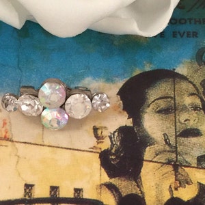 Puede incluir: Un grupo de cinco brillantes de cristal iridiscente transparente fijados en una base plateada. Los brillantes están dispuestos en una línea curva, con el brillante más grande en el centro.