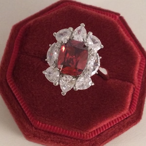May include: A red gemstone ring with a cluster of smaller clear gemstones surrounding the centre stone. The ring is displayed in a red velvet ring box.