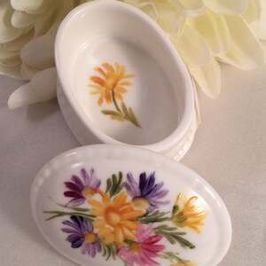 May include: White porcelain trinket box with a floral design. The lid features a bouquet of purple, pink, and yellow flowers. The base of the box has a single yellow flower.