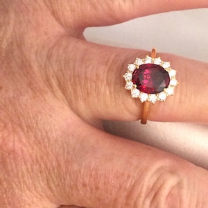 Puede incluir: Un anillo de oro con una piedra preciosa grande, de forma ovalada, de color rojo oscuro rodeada por un halo de piedras preciosas más pequeñas y transparentes.