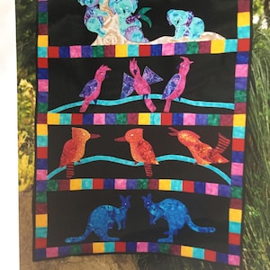 May include: A colorful quilt featuring Australian animals. Koalas, birds, and kangaroos are appliqued on a black background. The quilt has a border of colorful squares. The animals are in shades of blue, orange, and purple.