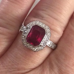 Può includere: Un anello d'argento con una grande pietra preziosa rossa a taglio smeraldo circondata da pietre preziose più piccole e trasparenti.