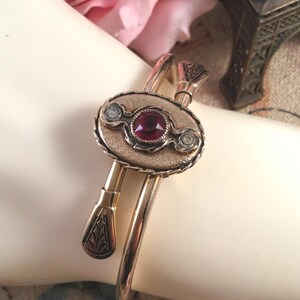 May include: Gold-toned bracelet with an oval centerpiece featuring a large, round, red gemstone and two smaller clear stones. The bracelet has decorative elements and a hinged design, showcasing a vintage aesthetic.