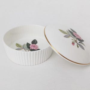 Könnte beinhalten: Weiße Keramik-Schmuckdose mit Deckel, verziert mit einem Rosenblütenmuster. Die Dose hat eine geriffelte Außenseite und einen goldenen Rand am Deckel. Der Deckel ist leicht geöffnet und zeigt das innere Blumenmuster.