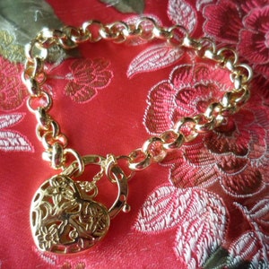 Könnte beinhalten: Ein goldenes Kettenarmband mit einem herzförmigen Schlossanhänger. Der Anhänger hat ein Blumenmuster und befindet sich auf einem roten Hintergrund.