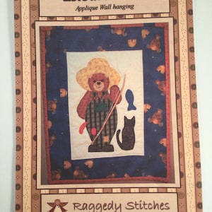 May include: A wall hanging pattern titled "Leisure Bear" featuring a brown bear in a straw hat and overalls, holding a fishing rod. A black cat and a blue fish are also depicted. The pattern is by Raggedy Stitches.