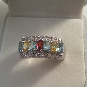 May include: A silver ring featuring a row of oval-cut gemstones in various colors, including blue, yellow, and red. The ring is encrusted with smaller, clear stones, adding sparkle. The ring is set against a light gray background.