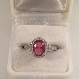 May include: A silver ring featuring a large oval pink gemstone encircled by smaller clear gemstones. The ring is displayed in a white ring box.