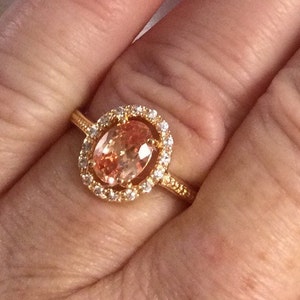 Puede incluir: Un anillo de oro con una gran piedra preciosa ovalada de color melocotón rodeada por un halo de piedras preciosas más pequeñas y transparentes.