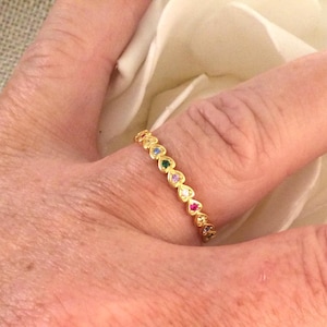 May include: A gold band ring with a row of small, colorful heart-shaped stones. The ring is on a finger.