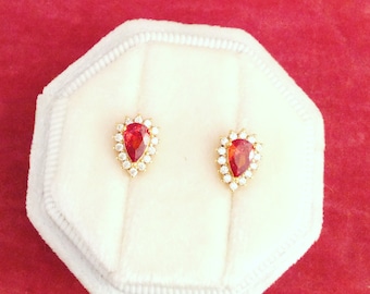 May include: A pair of gold stud earrings with pear-shaped, red gemstones surrounded by small, clear stones. The earrings are displayed in a white, octagonal jewelry box against a red background.
