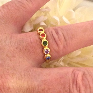 Puede incluir: Un anillo de oro con siete piedras redondas en un patrón de colores arcoíris. El anillo está en un dedo.