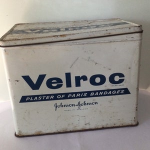 May include: A white metal tin with blue lettering that reads "Velroc Trade Mark Plaster of Paris Bandages Johnson & Johnson Made in England".