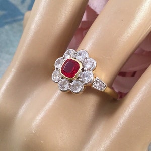 Op de afbeelding: Een gouden ring met een rode edelsteen omringd door kleinere witte edelstenen in de vorm van een bloem.