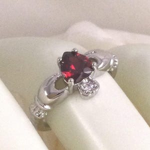 Puede incluir: Un anillo Claddagh plateado con una piedra preciosa roja oscura en forma de corazón. El anillo presenta dos manos sosteniendo una corona sobre el corazón y un pequeño grupo de piedras claras debajo. Diseño tradicional irlandés.