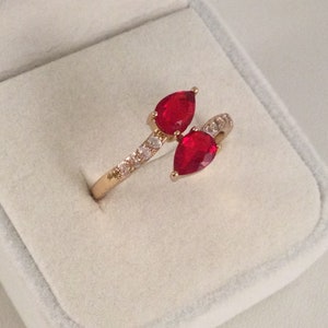 Könnte beinhalten: Ein goldener Ring mit zwei roten birnenförmigen Steinen und einer Reihe kleiner, klarer Steine.