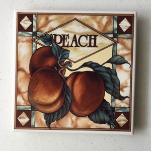 May include: A ceramic tile featuring a peach design. The tile depicts three peaches with green leaves and the word "PEACH" in a diamond-shaped frame.